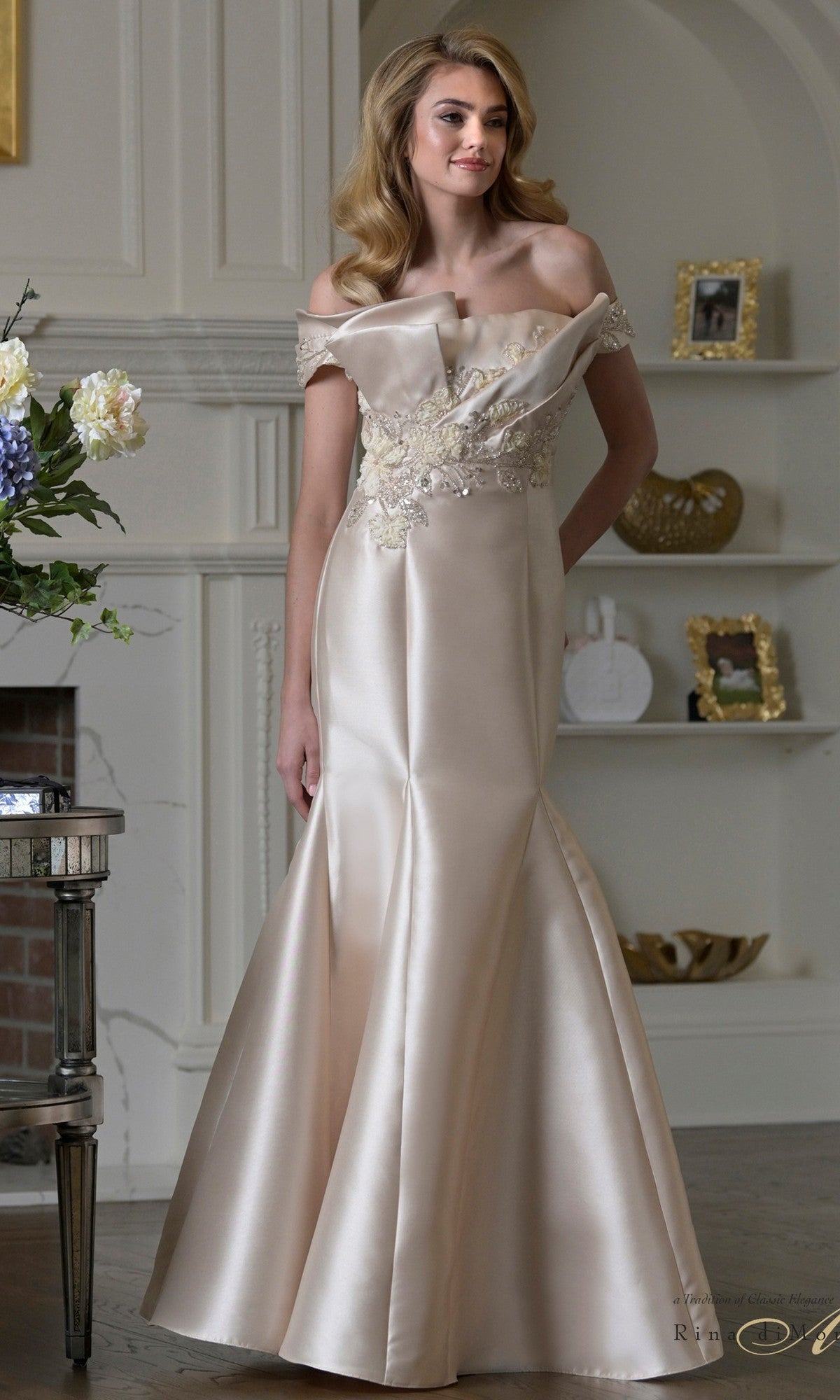 A woman stands indoors wearing the Rina Di Montella RD3408, a long champagne mermaid formal dress with off-the-shoulder sleeves and beaded floral appliques—an elegant choice for a mother of the bride.